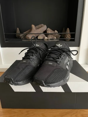 Svarta Adidas NMD sneakers - Säljer ett par svarta Adidas NMD sneakers med klassiska tre vita ränder på sidan och snygga detaljer på plösen och hälen. Skorna har snörning, platt sula och är tillverkade i syntet och mesh för en skön och modern känsla. 