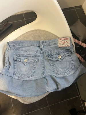 True Religion jeanskjol med volang - Säljer en ljusblå jeanskjol från True Religion, modell Joey Low Rise Flare. Kjolen har volang nedtill, klassiska jeansfickor både fram och bak samt synliga sömmar och knappdetaljer. Perfekt för dig som gillar street och denim. Jeans omsydda till kjol, lite slarvigt