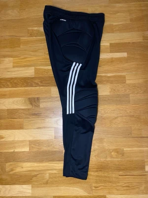 Svarta Adidas målvaktsbyxor M - Säljer ett par svarta Adidas målvaktsbyxor i storlek M. Byxorna har vadderade skydd vid knän och höfter samt klassiska vita ränder längs benen. Tillverkade i polyester och perfekta för fotbollsträning. Adidas-logga på låret.