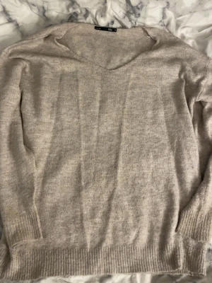 Beige stickad tröja från 157 - Mysig beige stickad tröja från 157 med v-ringning och långa ärmar. Tröjan har ribbade muddar vid ärmslut och nederkant, perfekt för lager på lager under kyliga dagar.