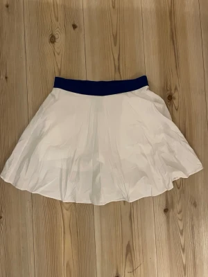 Vit golfkjol från J.Lindeberg - S - Säljer en snygg vit kortkjol från J.Lindeberg med blå midjeresår. Kjolen har en enkel och sportig design, perfekt för dig som gillar stilrena plagg. Tillverkad i polyester för en lätt och bekväm känsla. Liten blå logga nertill på kjolen.