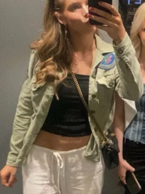 Grön jeansjacka med patch - Säljer en ljusgrön jeansjacka med cool patch på bröstet. Jackan har klassisk krage, knappar framtill och är croppad i modellen. Perfekt att slänga över en topp för en avslappnad look.