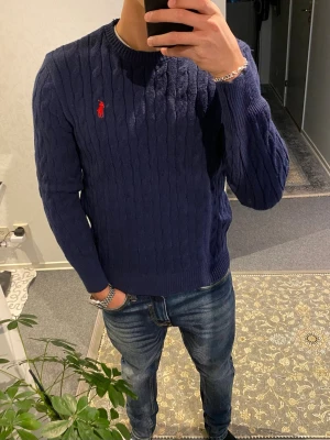 Mörkblå stickad tröja från Polo Ralph Lauren - Säljer en mörkblå stickad tröja från Polo Ralph Lauren med klassiskt kabelstickat mönster och röd broderad logga på bröstet. Tröjan har rund hals och långa ärmar med ribbade muddar. Perfekt för dig som gillar stilrena och tidlösa plagg.