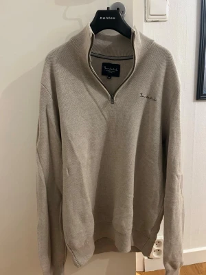 Beige stickad half zip-tröja Bondelid - Säljer en beige stickad tröja från Bondelid med half zip och hög krage. Tröjan har långärmade ribbade muddar och diskret broderad logga på bröstet. Perfekt för dig som gillar stilren och enkel design med lite extra edge.