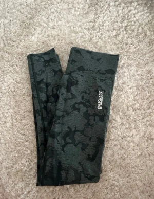 Mörkgröna camo tights från Gymshark - Säljer ett par snygga tights från Gymshark i mörkgrönt camo-mönster. De har hög midja och är tillverkade i ett stretchigt material som sitter skönt och följer kroppen. Perfekta för träning och gym. Gymshark-logga på benet + text vid höften. Köpta i strl XS, passar även S då de töjer sig lite vid användning.