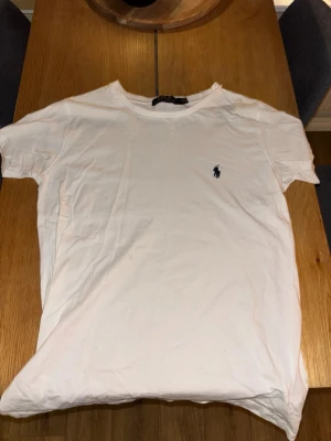 Vit t-shirt från Polo Ralph Lauren - Vit t-shirt från Polo Ralph Lauren i mjuk bomull. Klassisk design med korta ärmar och rund hals. Diskret blå logga broderad på bröstet. Perfekt för en clean och stilren look.