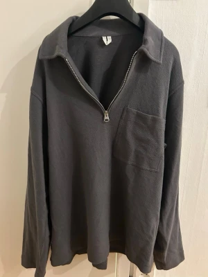 Mörkgrå half zip tröja med krage - Mörkgrå långärmad tröja med stor krage och half zip-dragkedja framtill. Tröjan har en bröstficka på vänster sida och är gjord i mjukt bomullstyg. Passar perfekt för chill dagar och har en relaxed vibe.