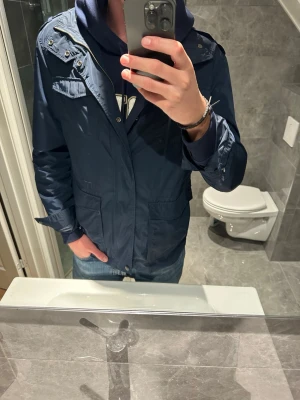 Marinblå vindjacka från Gant, M - Säljer en marinblå vindjacka från Gant i storlek M. Jackan har flera fickor framtill, axelklaffar och dragkedja med tryckknappar. Jag på bilden är 188cm och den sitter bra på mig. Hör av er vid funderingar;)