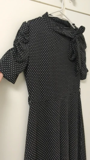 Svart prickig klänning Voodoo Vixen - Passform S. Svart kortklänning från Voodoo Vixen med vita prickar, puffiga kortärmar och en söt knyt-detalj vid halsen. Klänningen har en mjuk, följsam passform och är tillverkad i ett stretchigt syntetmaterial. Perfekt för dig som gillar retrostil och vill sticka ut.