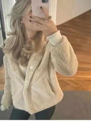 Beige teddyjacka med huva - Supermjuk och fluffig teddyjacka i beige med huva och fickor på sidorna. Jackan har en avslappnad passform och stängs framtill. Perfekt för dig som vill ha en mysig jacka under kyligare dagar. Ens fickan han en tråd som har lossnat så att en del flärpas ut men det syns inte när man har på sig jackan och man kan vara vika in det. Hör av er vid fler frågor💗💗