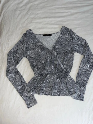 Grå omlottopp med paisleymönster Bik Bok - Säljer en grå omlottopp från Bik Bok med paisleyinspirerat mönster i svart och vitt. Toppen har lång ärm, v-ringad hals och markerad midja som ger en snygg siluett. Perfekt att styla med jeans eller kjol för en cool look.