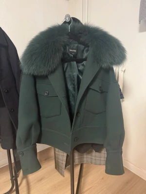 Mörkgrön pälsjacka från Meotine XS/S - Snygg mörkgrön jacka från Meotine med stor fluffig päls på kragen. Jackan har en croppad passform, knappar framtill och två fickor med lock. Materialet är ullblandning och pälsdetaljen ger en lyxig vibe. Använd ett fåtal gånger och inga defekter