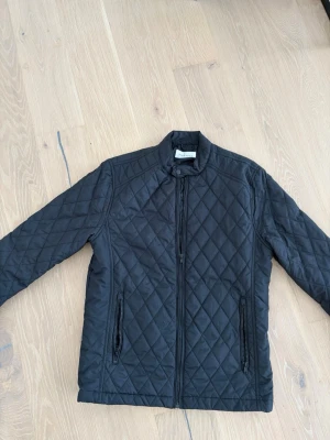 Svart quiltad jacka - Snygg svart quiltad jacka från pier one med dragkedja framtill och två fickor med dragkedja. Jackan har en stilren ståkrage och ett rutigt mönster över hela ytan. Perfekt för dig som gillar en clean och modern look. Storlek medium