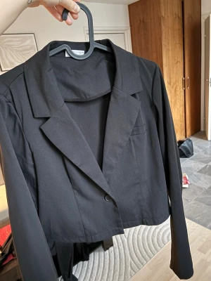 Svart croppad kavaj - Snygg svart croppad kavaj med klassisk krage och en knapp framtill. Jackan har lång ärm och enkel, stilren design som passar till många olika looks. Perfekt för dig som vill ha en modern twist på den klassiska kavajen.