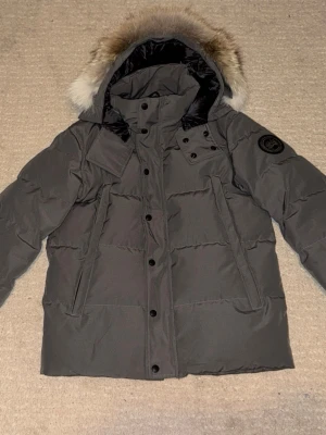 Grå dunjacka från Canada Goose - Säljer en grå dunjacka från Canada Goose med svart logotyp på ärmen och avtagbar huva med päls. Jackan har långa ärmar, flera fickor och stängs med både dragkedja och knappar. Perfekt för kalla vinterdagar.