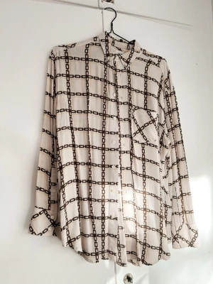 Hope skjorta med kedjemönster - Säljer en beige skjorta med brunt och svart kedjemönster och knappar framtill. Skjortan har lång ärm, klassisk krage och en bröstficka. Materialet känns mjukt och flowy, 100% viskos. Den har ett litet hål på baksidan och några svaga fläckar. Rymlig storlek. 