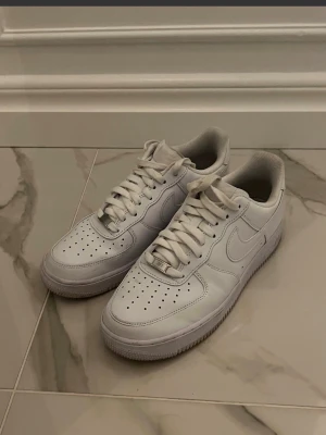 Nike Air Force 1 vita sneakers - Säljer ett par klassiska Nike Air Force 1 sneakers i helvitt skinn med snörning och perforerad tå. Ikonisk siluett med rund tå och platt sula, perfekt för dig som gillar stilrena och tidlösa sneakers. Passar till alla dina outfits och är superbekväma.