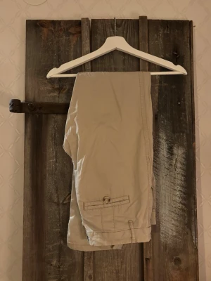 Beige chinos från 365 Sunshine, stl. 36 - Snygga beige chinos från 365 Sunshine med klassisk passform och knappdetalj vid bakfickan. Byxorna har bälteshällor och är gjorda i mjuk bomull som är skön att ha på sig. Perfekta för dig som gillar en clean och enkel stil.