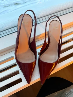 Vinröda pumps med spetsig tå från MNG - Säljer ett par eleganta vinröda pumps från MNG med spetsig tå och smal klack. Skorna har en snygg slingback-design och är tillverkade i glansigt syntetmaterial som ger en lyxig känsla. Perfekt för dig som gillar stilrena och trendiga skor.  Det finns två skavanker på skorna (se bild) ❤️