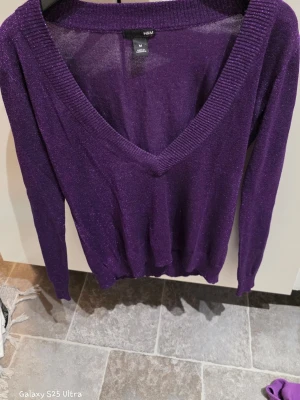 Glittrig lila v-ringad tröja H&M - Säljer en snygg lila tröja från H&M i storlek M. Tröjan har glittrigt material, djup v-ringning och långa ärmar. Tillverkad i viskos och polyester, perfekt för dig som gillar att sticka ut med lite extra shimmer.
