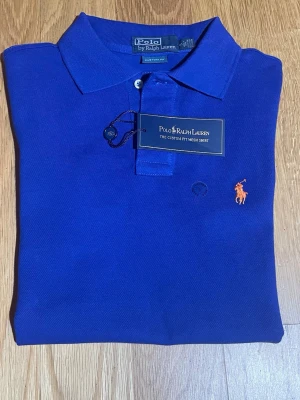 Blå pikétröja från Polo Ralph Lauren - Säljer en blå pikétröja från Polo Ralph Lauren i storlek S. Tröjan har klassisk krage, korta ärmar och två knappar framtill. Den är gjord i bomull och har den ikoniska orangea Polo-loggan broderad på bröstet. Custom fit-modell med stilren look.