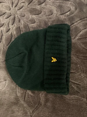 Grön stickad mössa med broderad detalj - Snygg mörkgrön stickad mössa från Lyle & Scott med gul broderad logga framtill. Mössan har bred uppvikt kant och är tillverkad i mjukt material som håller dig varm. Perfekt för kalla dagar och enkel att matcha med din jacka.