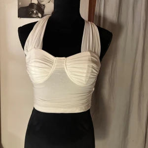 Vit korsettinspirerad topp - Säljer en vit korsettinspirerad topp med breda axelband och draperad framsida. Toppen har en oneshoulder-liknande design baktill och är croppad. Materialet känns stretchigt och mjukt, perfekt för att skapa en cool och trendig look.