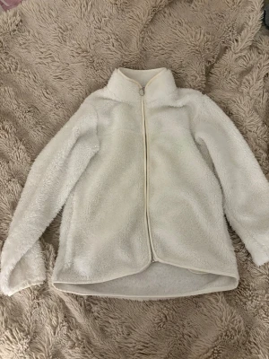 Vit teddyjacka med dragkedja - Mysig vit teddyjacka med hög krage och hel dragkedja framtill. Jackan har långärmad design och är gjord i fluffigt fleece-material som håller dig varm under kyliga dagar. Perfekt för lager på lager och enkel att matcha med olika outfits.