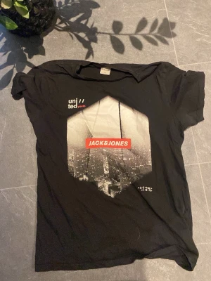 Svart t-shirt Jack & Jones Core - Svart t-shirt från Jack & Jones Core med tryck framtill som visar en stadssilhuett och texten 'united youth' samt röd logga. Klassisk rund hals och korta ärmar. Materialet är mjuk bomull, perfekt för chill dagar. Passar M-L 