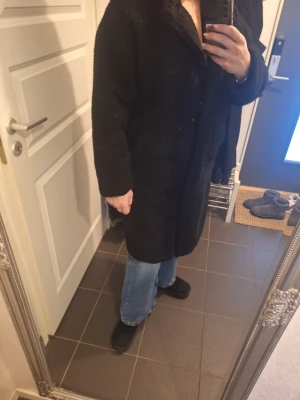 Svart teddyjacka i oversized modell - Mysig svart teddyjacka med lång ärm och oversized passform. Jackan är knälång och har en fluffig, mjuk yta som ger en cool och avslappnad vibe. Perfekt för dig som gillar streetstyle och vill hålla dig varm under höst och vinter.