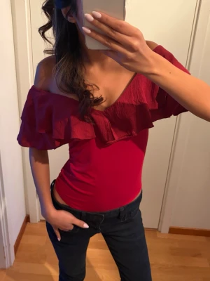 Röd body med volang - Säljer en snygg röd body med omlott-design och bred volang längs axlarna. Toppen är offshoulder och har en v-ringad hals. Materialet känns mjukt och lätt, perfekt för att ge en cool vibe till din outfit.