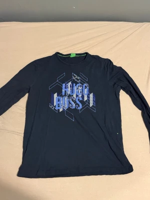 Mörkblå långärmad t-shirt Hugo Boss - tröja  - Snygg mörkblå långärmad t-shirt från Hugo Boss med coolt grafiskt tryck i blått och vitt på bröstet. Rund hals och mjukt material som känns skönt mot huden. Perfekt för dig som gillar stilrena märkesplagg med en modern touch.