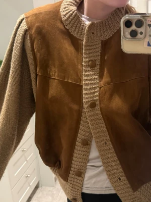 Beige och brun stickad bomberjacka - Säljer en unik bomberjacka i beige och brun med stickade ärmar, mocka och muddar. Framsidan och ryggen är i mocka och ärmarna är stickade. Jackan har knappar framtill och en avslappnad passform. Perfekt cool vår/höst jacka. Använd ca 2/3 gånger.