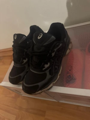 Svarta Asics GEL sneakers - Svarta Asics GEL sneakers med grå detaljer och klassisk snörning. Skorna har en rund tå och platt sula, med tydlig Asics-logga på sidan och GEL-tryck bak. Materialet är en mix av syntet och mesh för bra ventilation och komfort. Perfekta för dig som gillar sportig stil.