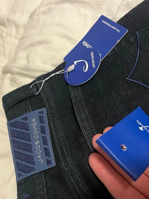 Jacob Cohën mörkblå jeansbyxor - Säljer ett par mörkblå jeansbyxor från Jacob Cohën med snygga blå detaljer och klassisk femficksdesign. Byxorna har exklusiva metallknappar och patch bak i blått. Materialet är jeans med en lyxig känsla och passformen är normal.