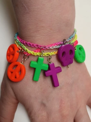 Färgglad flätad charm-armband - Säljer ett riktigt färgstarkt wrap armband med flätad syntettråd i neonfärger och stora plastberlocker i form av peace-tecken, kors och döskalle. Armbandet har orange, gröna, lila och rosa detaljer som verkligen poppar och ger en lekfull vibe.