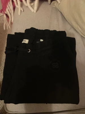 Svarta mjukisbyxor från Sandro - Svarta mjukisbyxor från Sandro med dragsko i midjan och diskret logga på benet. Byxorna har en relaxed passform och är tillverkade i mjukt material som känns skönt mot huden. Perfekta för chill dagar eller när du vill vara bekväm men ändå se snygg ut.