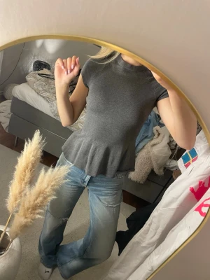 Mörkgrå peplum topp med kort ärm - Säljer en mörkgrå peplum topp med korta ärmar och rundad hals. Toppen har en volangkant nedtill som ger en snygg siluett. Materialet är mjukt och stretchigt, troligtvis bomull eller polyester. Perfekt att styla med jeans eller kjol.