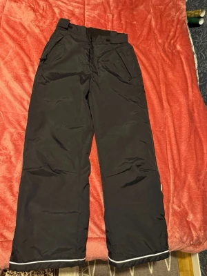 Svarta täckbyxor med reflexdetalj - Svarta täckbyxor med bred passform och reflexdetalj längst ner vid bensluten. Byxorna har praktiska fickor framtill och är gjorda i ett slitstarkt syntetmaterial som passar perfekt för kalla dagar. Midjan är justerbar med kardborreband för extra komfort. Den har använts ca 2 gånger. Den helt ny och ren. Storlek 152