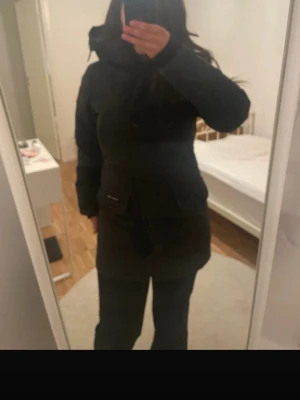 Svart jacka från Canada Goose - Säljer en svart jacka från Canada Goose med huva och klassisk logga på ärmen. Jackan är lång, har stora fickor framtill och stängs med både dragkedja och knappar. Perfekt för kalla vinterdagar och har en clean, stilren look.