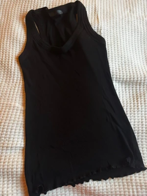 Svart ribbad linnetopp - Säljer en svart ribbad linnetopp med rundad hals och ärmlös design. Toppen har en enkel och stilren look med lätt vågig kant nedtill. Perfekt att matcha med jeans eller kjol för en chill och snygg outfit.