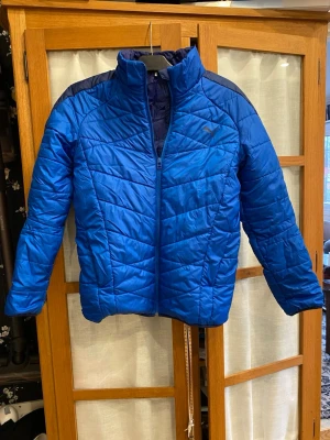 Blå quiltad pufferjacka från Puma - Blå pufferjacka från Puma med quiltad design och hög krage. Jackan har dragkedja framtill och mörkblå detaljer vid axlarna. Perfekt för kyliga dagar och har en sportig look som passar dig som gillar att sticka ut.