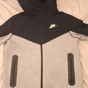 Grå och svart huvtröja från Nike - Säljer en snygg grå och svart huvtröja från Nike med dragkedja och huva. Tröjan har två praktiska fickor med dragkedjor framtill och Nike-logga på bröstet. Perfekt för en sportig look!