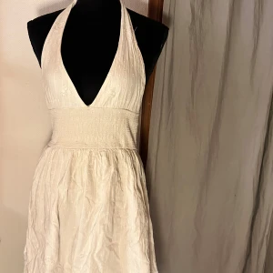 Vit omlottklänning med halterneck - Säljer en vit kortklänning med halterneck och omlott-detalj. Klänningen har djup v-ringning, smockad midja och ett diskret prickigt mönster. Materialet känns lätt och luftigt, perfekt för varma dagar. Knytband i nacken och mjukt fallande kjol.