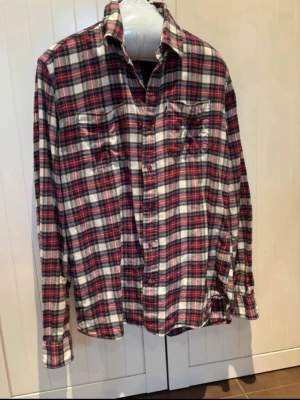 Rutig Tommy Hilfiger skjorta i bomull - Riktigt snygg rutig skjorta från Tommy Hilfiger i mjuk bomull. Skjortan har klassiskt röd, blå och vit rutmönster, två bröstfickor med knapp och diskret logga på ena fickan. Perfekt oversized fit och långa ärmar med knappar vid manschetten.