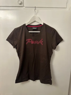 Brun Peak Performance t-shirt - Snygg brun t-shirt från Peak Performance med rosa broderad logga på bröstet. Klassisk rund halsringning och korta ärmar. Tillverkad i mjuk bomull som känns skön mot huden. Perfekt för dig som gillar sportiga och stilrena plagg.
