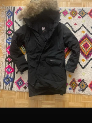 Svart Canada Goose parkajacka M - Svart parkajacka från Canada Goose med fluffig päls på huvan och flera fickor framtill. Jackan är fodrad med dun och har dragkedja samt knappar. Materialet är polyester och bomull på utsidan, med nylonfoder. Perfekt för kalla vinterdagar.