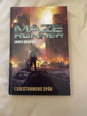 Maze Runner: I solstormens spår - En spännande bok med dramatiskt omslag där ungdomar står mitt i en stad i kaos, omgivna av eld och förstörda bilar. Perfekt för dig som gillar action, mysterier och dystopiska äventyr. Passar unga vuxna som söker nervkittlande läsning.