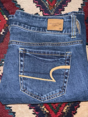 Bootcut jeans från American Eagle - Snygga blå bootcut jeans från American Eagle med stretch. Klassisk femficksdesign och coola kontrastsömmar på bakfickorna. Jeansen har låg midja och två kopparfärgade knappar fram. Perfekt passform och mjukt denimtyg.
