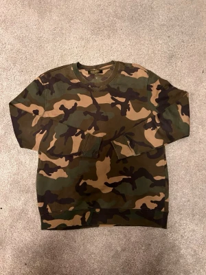 Camouflage sweatshirt från Valentino - Säljer en grön och brun camouflage-mönstrad sweatshirt från Valeytino. Tröjan har rund hals, ribbade muddar och en liten nitar-detalj bak i nacken. Perfekt för dig som gillar streetwear och vill sticka ut med coolt mönster. Obs stolek L men passar M